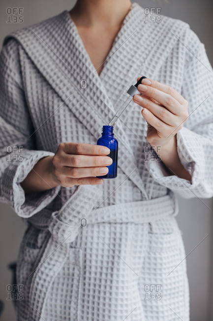 Hands of unrecognizable woman in bathrobe holding bottle of cosmetic serum and a pipette.
