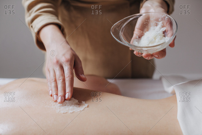 Hands of unrecognizable woman masseuse applying skin scrub on client's back.