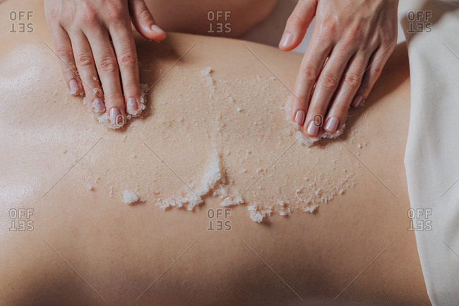 Hands of unrecognizable woman masseuse applying skin scrub on client's back.