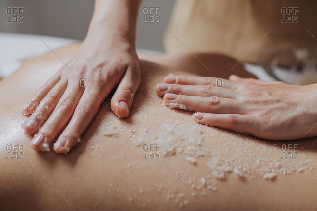 Hands of unrecognizable woman masseuse applying skin scrub on client's back.