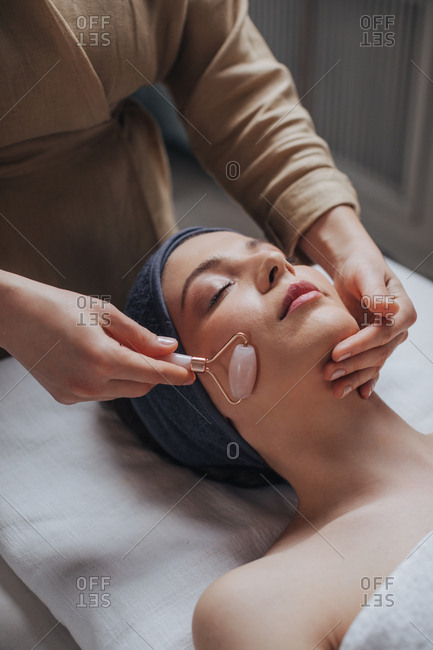 Beautiful young woman enjoying face massage with crystal roller at spa salon.