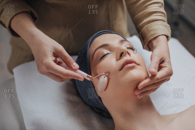 Beautiful young woman enjoying face massage with crystal roller at spa salon.