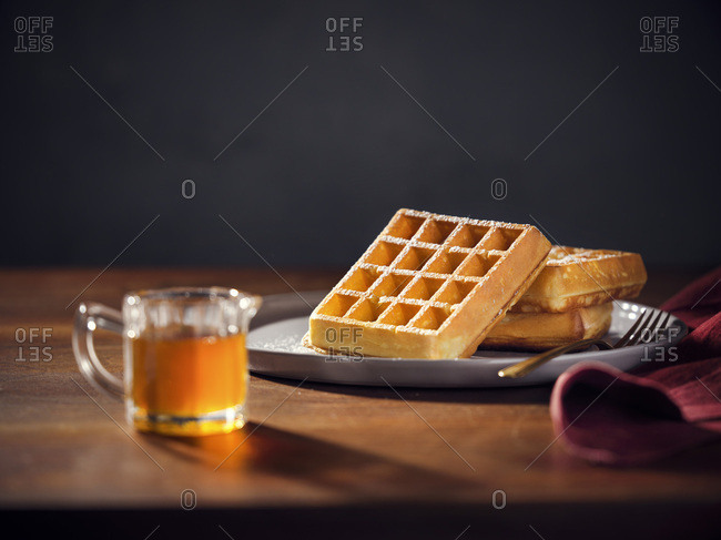 A plate with 3 belgian waffles with powdered sugar. Pitcher of maple syrup in the foreground. Warm, moody lighting on wood tabletop.