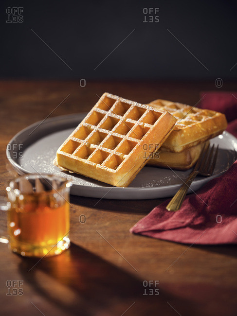 A plate with 3 belgian waffles with powdered sugar. Pitcher of maple syrup in the foreground. Warm, moody lighting on wood tabletop.