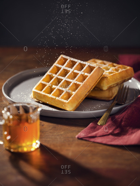 A plate with 3 belgian waffles being sprinkled with powdered sugar. Pitcher of maple syrup in foreground. Warm, moody lighting on wood tabletop.
