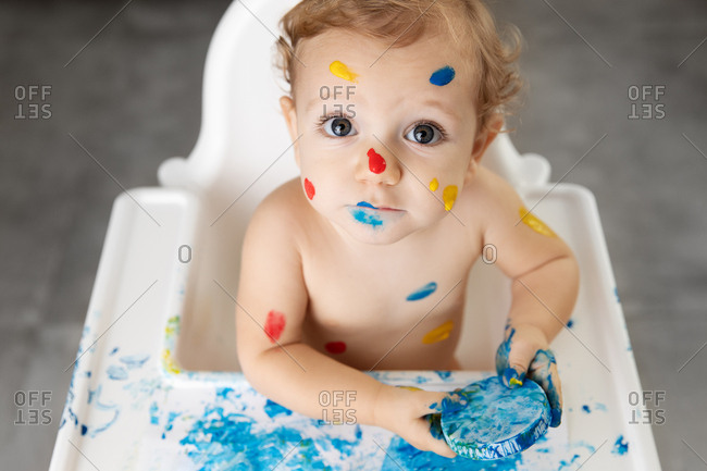 Baby in high chair with finger paint on face and body looking up
