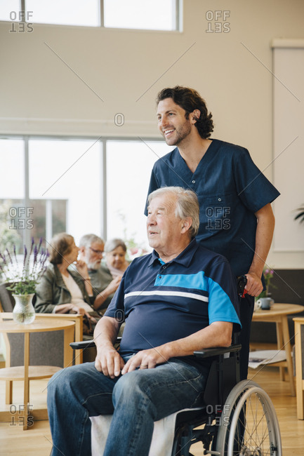 Smiling male nurse pushing disabled senior man on wheelchair at retirement home