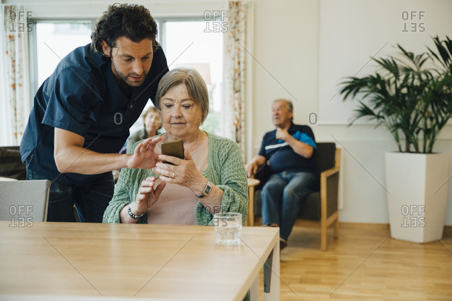 Confident male caregiver assisting senior woman using smart phone in nursing home