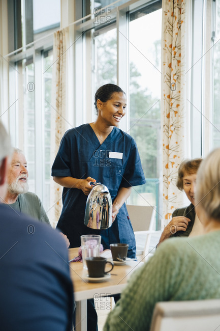 Smiling female nurse serving drink to senior women and men at dining table in nursing home