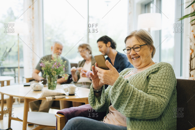 Smiling retired senior woman using smart phone while sitting on chair with friends and caregiver in background