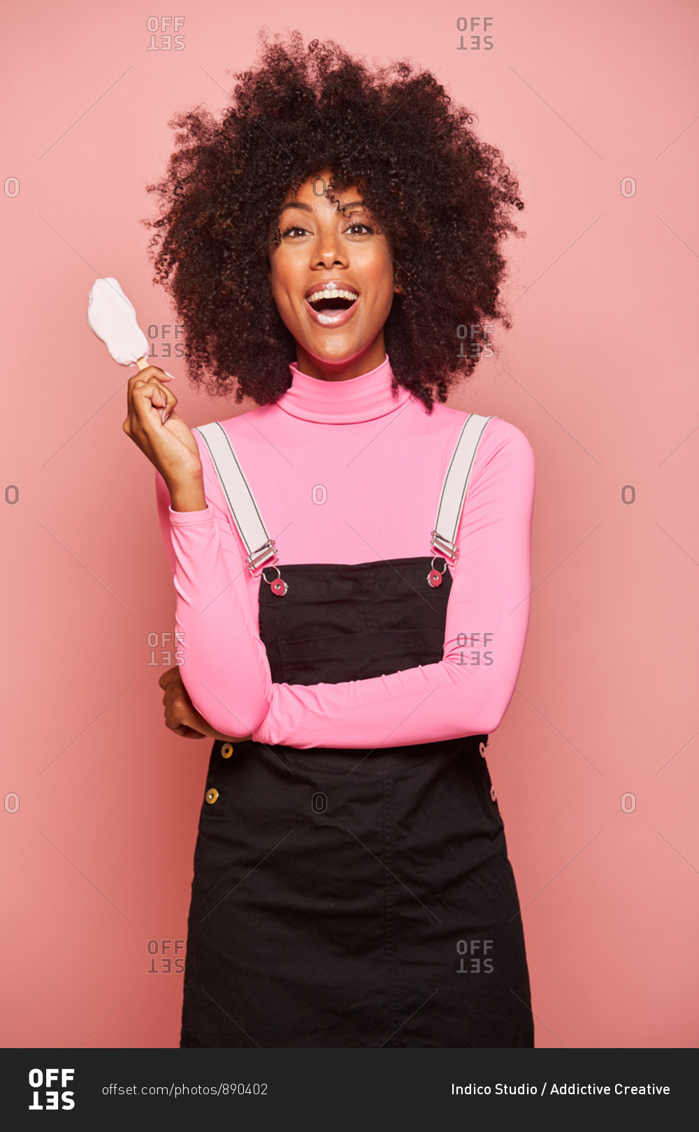 Positive excited young African American female with smeared lips wearing pink turtleneck and black overall holding ice cream on stick and looking at camera while standing against pink background