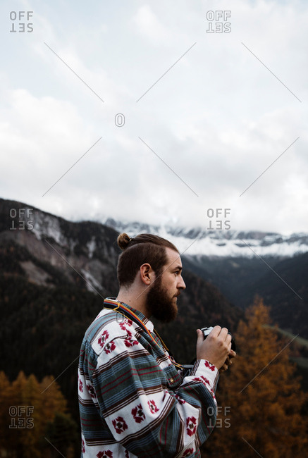 Side view of man traveler in casual wear standing with professional camera delighting in view of forest with Dolomites mountains on background at Italy