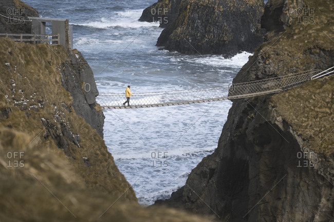 From above side view of traveler passing over Carric a Rede rope bridge suspended between rocky cliffs and sea waves crashing on rocks in background