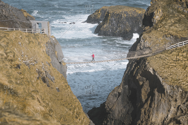 From above side view of traveler passing over Carric a Rede rope bridge suspended between rocky cliffs and sea waves crashing on rocks in background
