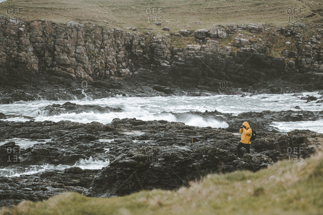 Male traveler standing on rock with camera on tripod and taking picture of seascape on cloudy gloomy day on Northern Ireland coastline
