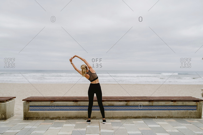 Back view of anonymous female athlete in active wear standing with raised hands in in lock and stretching near seashore on cloudy weather