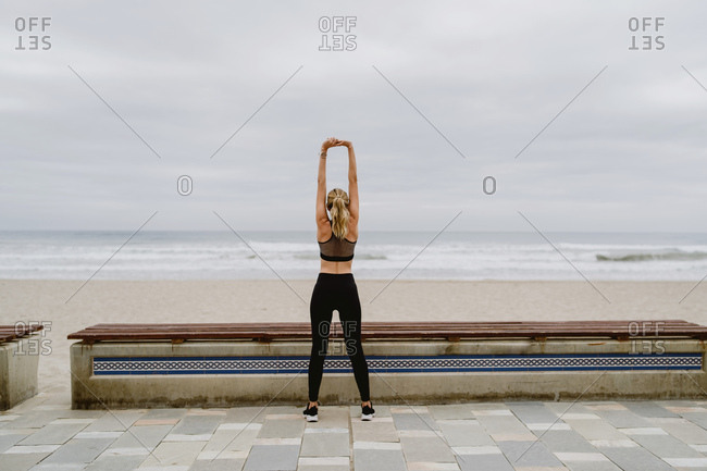 Back view of anonymous female athlete in active wear standing with raised hands in in lock and stretching near seashore on cloudy weather