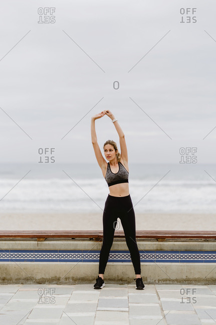 Athlete woman in active wear standing with raised hands in in lock and stretching near seashore on cloudy weather