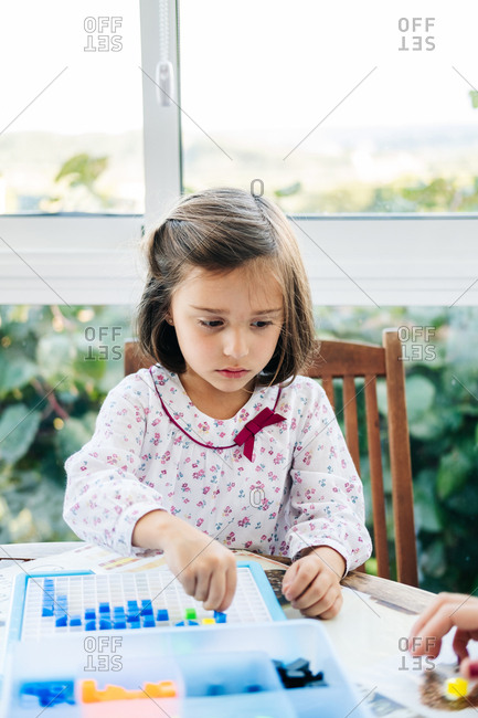 Adorable concentrated little girl putting small colorful blocks to board and creating mosaic while playing educational game