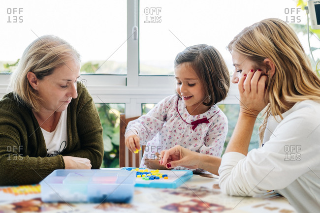 Middle aged woman with little girl and adult daughter having fun and playing board game creating picture with colorful mosaic pieces while sitting at round table on terrace