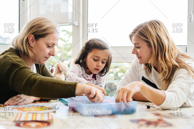 Middle aged woman with little girl and adult daughter having fun and playing board game creating picture with colorful mosaic pieces while sitting at round table on terrace