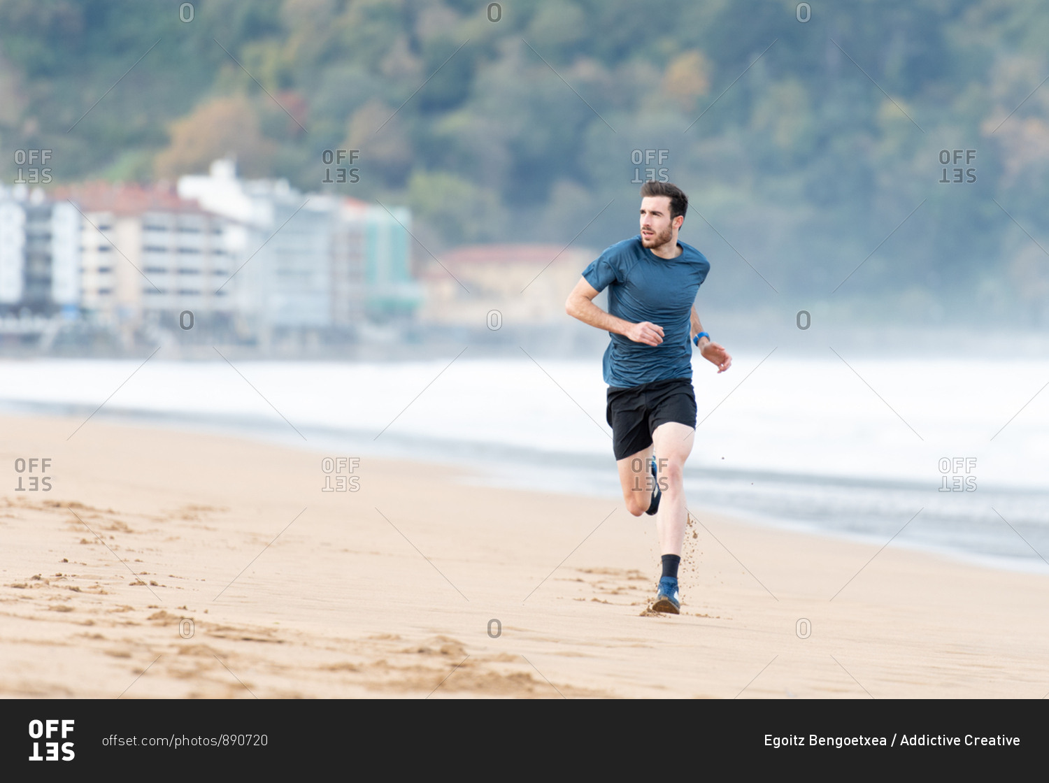 Bearded male athlete in active wear running during empty sandy beach with green mountains on blurred background