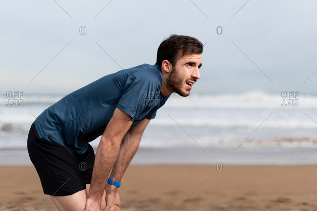 Side view of bearded sporty man in active wear taking a break after a long run on sandy empty seaside and looking away