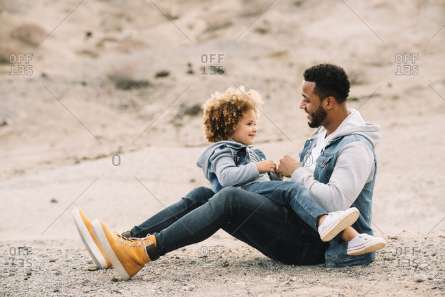 Cheerful bearded ethnic man dressed in stylish clothes lying on back with legs bent and holding curly happy ethnic toddler on bright sandy hills at daytime