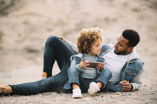 African American bearded casual man lying on sandy ground leaning on elbow and bending leg at knee looking at curly ethnic toddler in denim clothes sitting beside with mobile phone