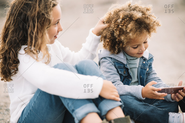 Smiling casual curly haired child resting on ground with crossed legs holding mobile phone while content girl sitting clasping knees and touching hair of friend with interest