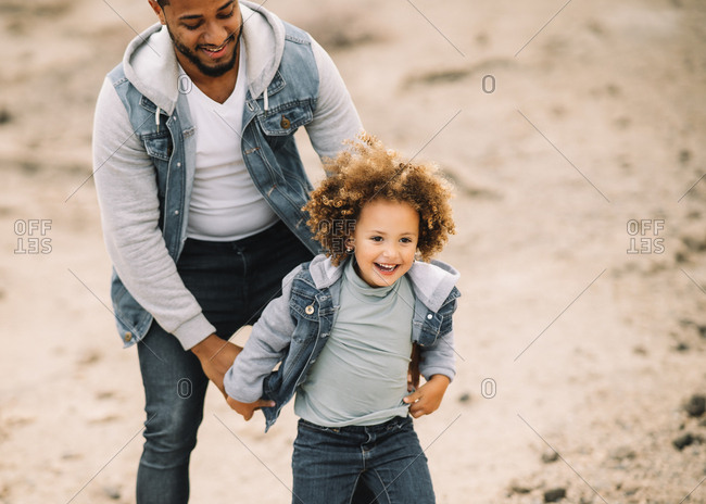 Cheerful bearded ethnic man dressed in stylish clothes holding playing with curly happy ethnic toddler on bright sandy hills at daytime