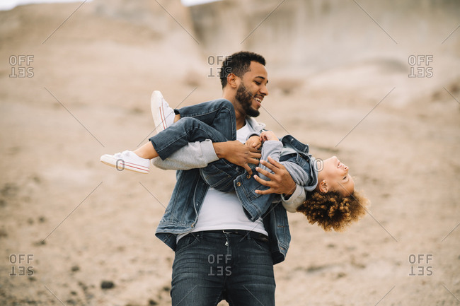 Cheerful bearded ethnic man dressed in stylish clothes holding playing with curly happy ethnic toddler on bright sandy hills at daytime