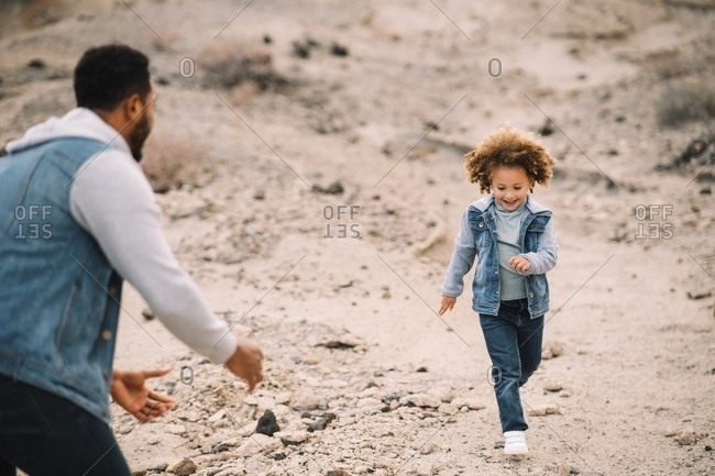 Cheerful bearded ethnic man dressed in stylish clothes playing with curly happy ethnic toddler on bright sandy hills at daytime