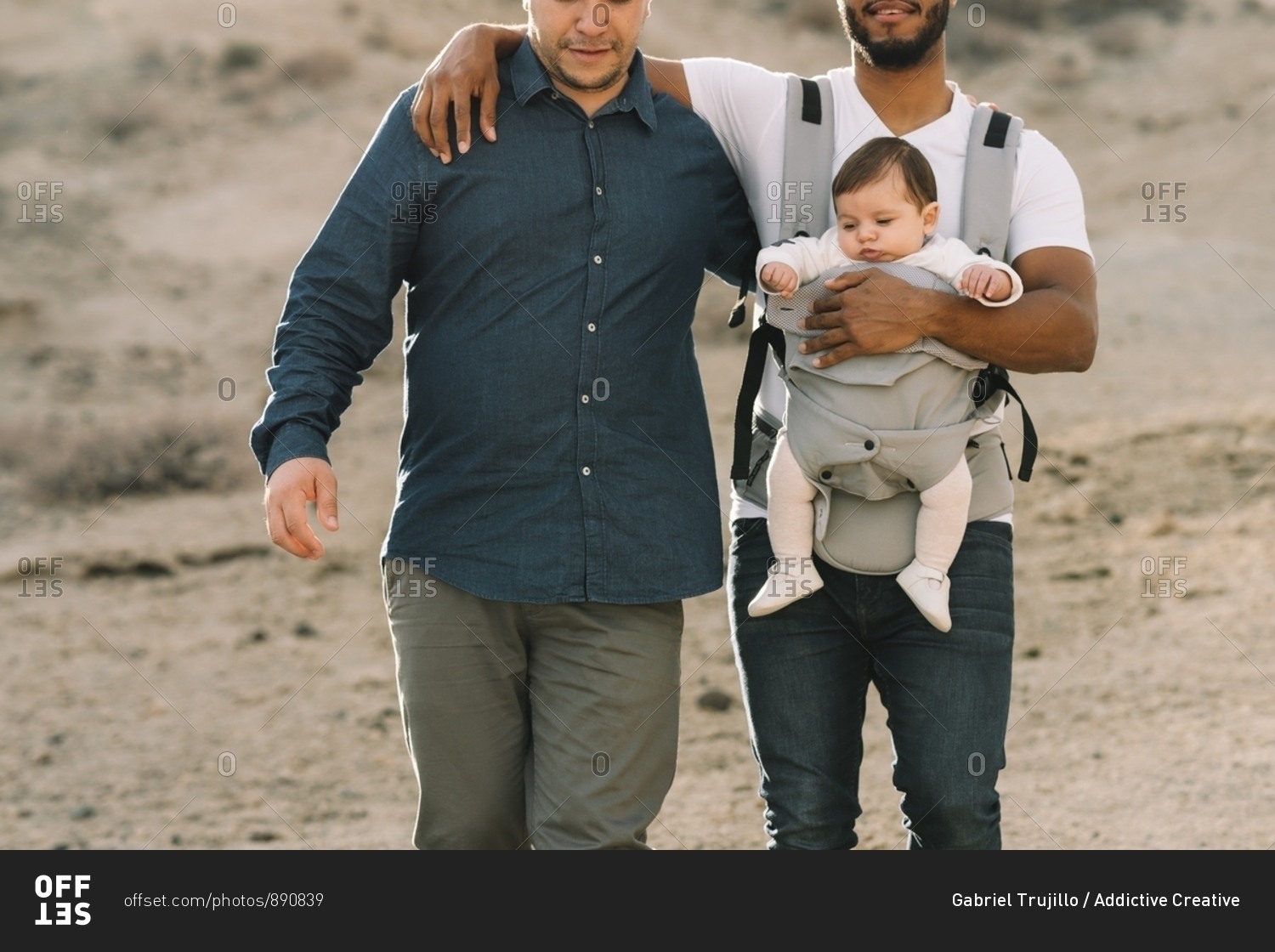 Crop black casual man holding on shoulder anonymous male boyfriend and holding little calm baby in grey carrier while strolling on nature at daytime