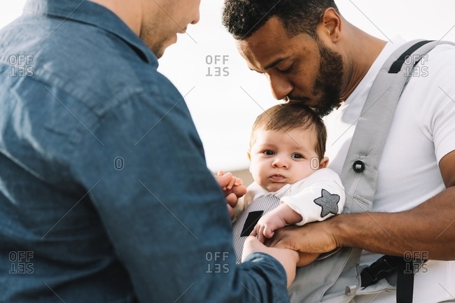 Side view of black casual man holding hand of anonymous male boyfriend and holding little calm baby in grey carrier while strolling on nature at daytime