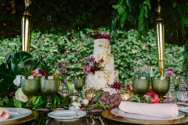 White and gold tiered cake with flowers placed on wedding table with plate and glasses decorated with fruits and flowers against green trees