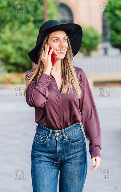 Content long haired lady in fashionable black hat and shirt walking on the street while calling on mobile phone and looking away at city street
