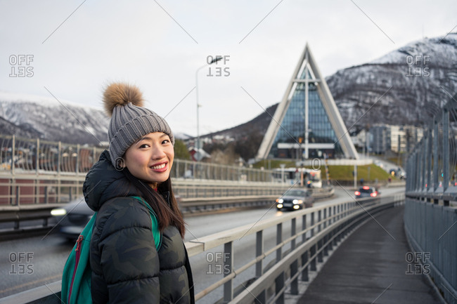 Attractive adult Asian woman in warm clothes with backpack smiling at camera while standing on street against blurred exterior of amazing triangle shaped church and snowy hills in Norway