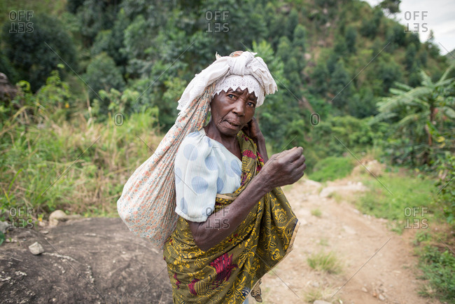 Uganda - November, 26 2016: Full body senior African lady looking at camera and carrying sack on back while walking on path in forest