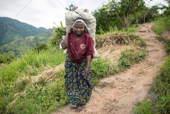 Uganda - November, 26 2016: Full body senior African lady looking at camera and carrying sack on back while walking on path in forest