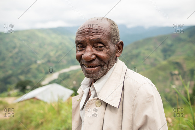 Uganda - November 26, 2016: Pensive senior African man in casual clothes looking at camera with kind gaze against amazing blurred scenery of green forested hills in daytime
