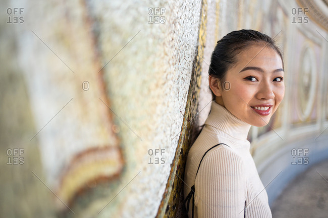 Side view of content Asian female on vacation in beige roll neck leaning on wall with fresco while smiling and looking at camera at St Peters Basilica in Vatican at Rome
