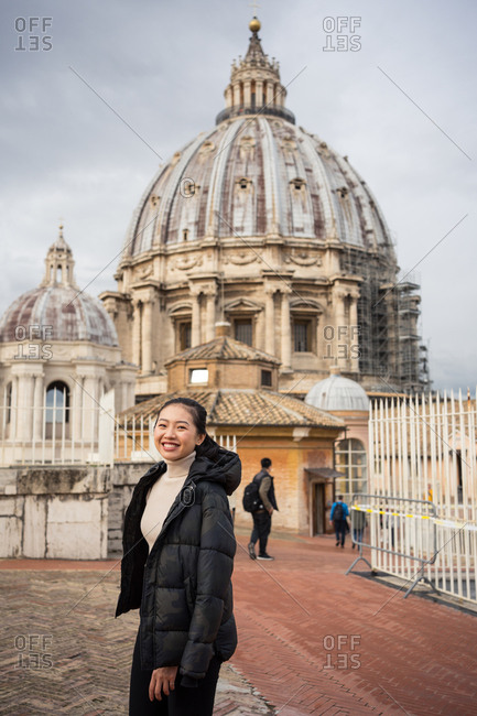 Pleased Asian female tourist in warm clothing smiling and looking at camera with beautiful ancient St Peters basilica in Vatican at Rome on background