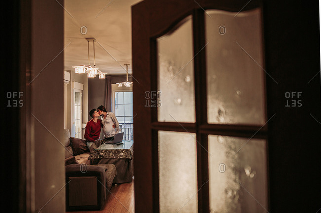 Young man in red jumper sitting on couch and kissing pregnant wife standing nearby while spending free time at home and enjoying time together