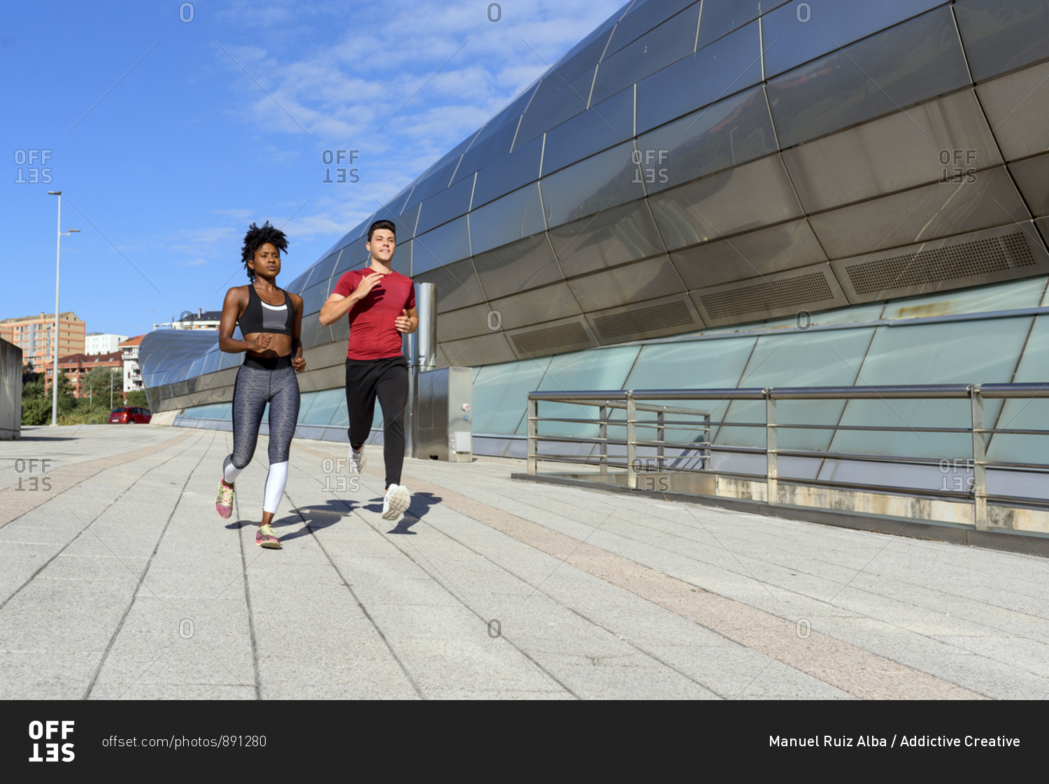 Young athletic African American female in top and leggings jogging with sportive male friend next to modern city construction in summer day