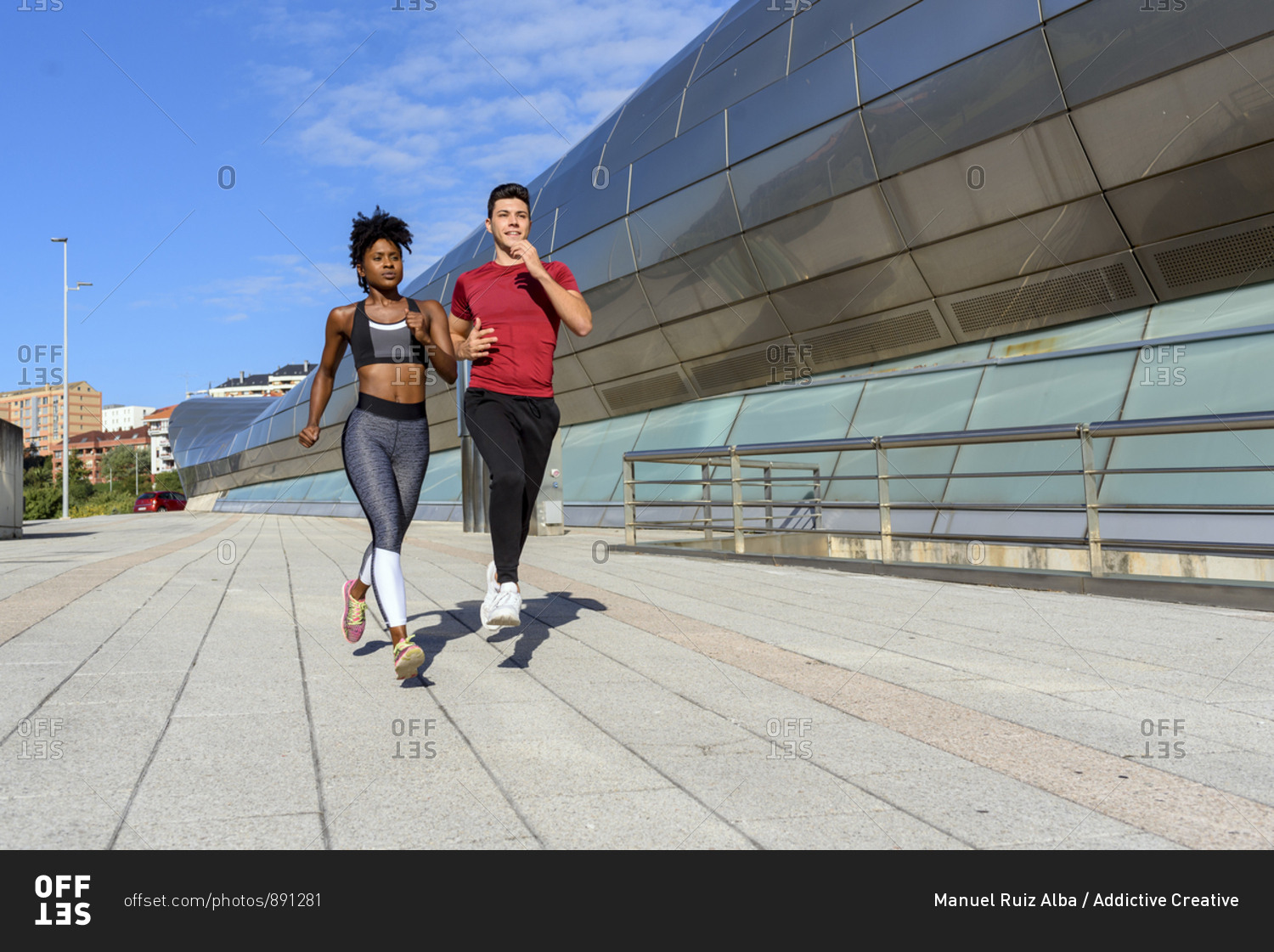 Young athletic African American female in top and leggings jogging with sportive male friend next to modern city construction in summer day