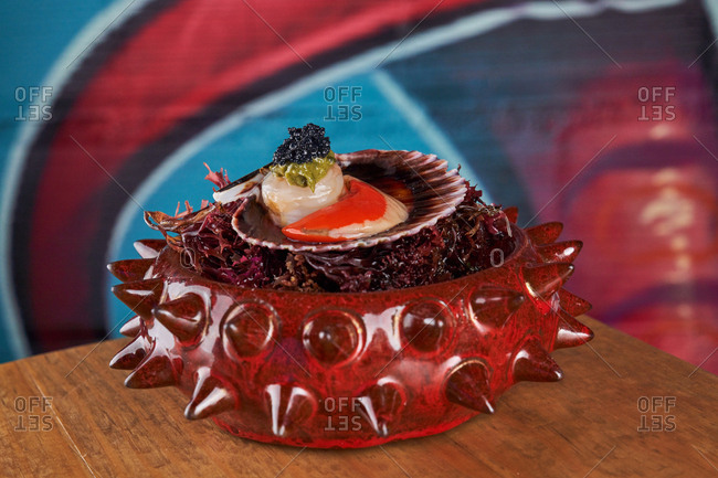 Served delicatessen in small oyster shell on modern shiny ceramic spiked bowl on wooden table in restaurant