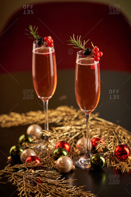 Pair of glasses of red cocktail decorated with berries and green herbs placed on black surface with green and gold tinsel and Christmas balls against blurred gold background