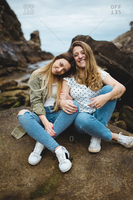 Relaxed women sitting on stone on hill and hugging while resting after walking in countryside on cloudy spring day and enjoying time together on cloudy spring day