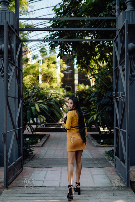 Back view of fashion trendy Asian woman with long dark hair in yellow short dress walking along metal decorative fence and looking at camera over shoulder
